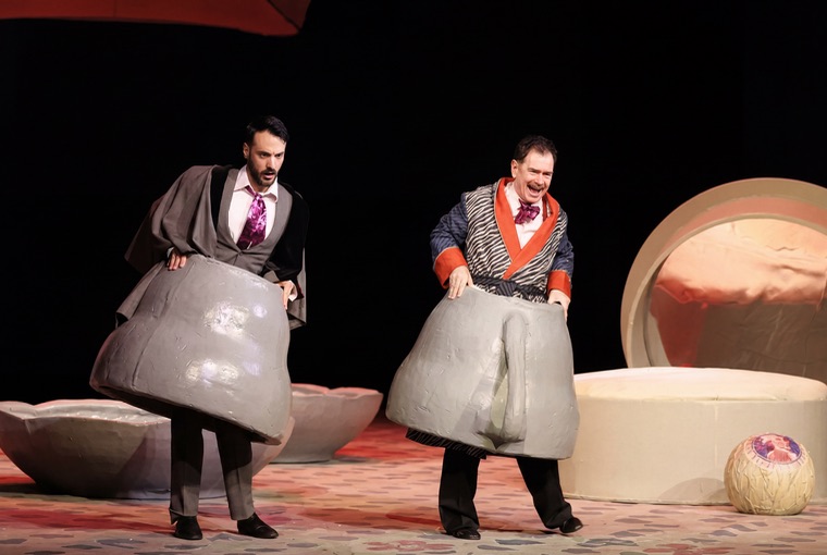 Two performers in comic costumes carrying bathtubs on stage